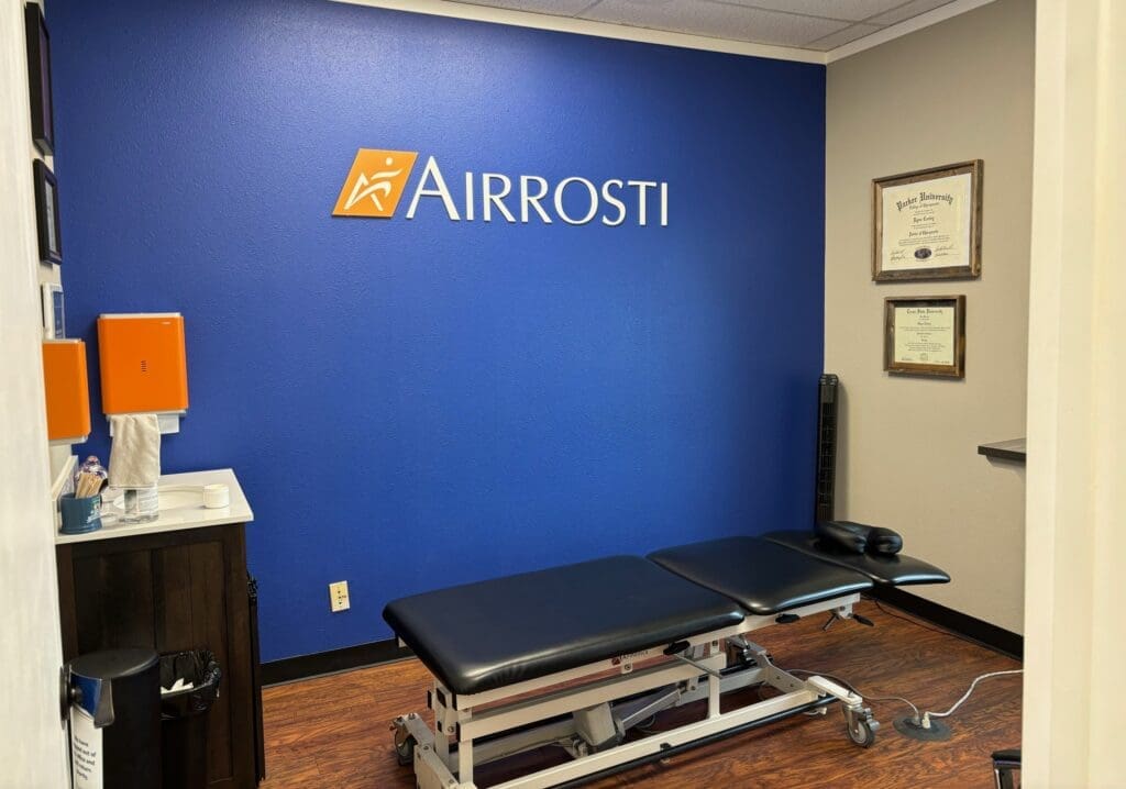 a photo of the treatment room inside Airrosti Rockwall. the treatment table is located center of the room and there is an Airrosti logo printed on the back wall.